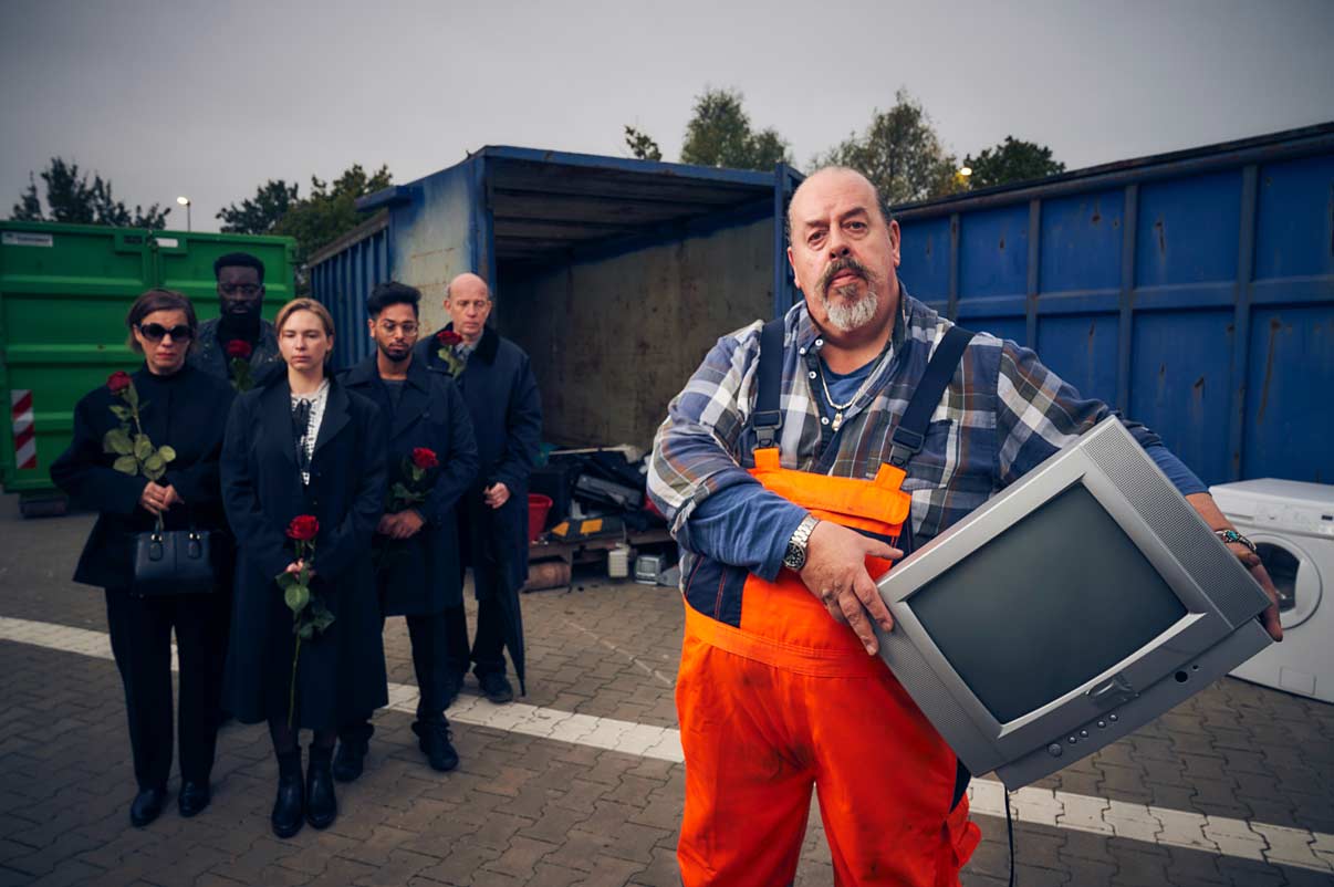 Wertstoffhofarbeiter hält entsorgten Fernseher, während eine Gruppe von Menschen im Hintergrund, mit Rosen in den Händen, um das Gerät trauert.